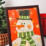 Decorate your home for the holiday season with this easy-to-make Lighted Snowman Picture. It’s fun to put together and perfect for hanging up around the home to get into the holiday spirit. This DIY Christmas craft is made with a gift bag and a dollar store frame. It is a fun dollar store Christmas craft.