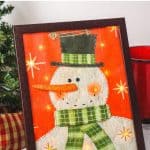 Decorate your home for the holiday season with this easy-to-make Lighted Snowman Picture. It’s fun to put together and perfect for hanging up around the home to get into the holiday spirit. This DIY Christmas craft is made with a gift bag and a dollar store frame. It is a fun dollar store Christmas craft.